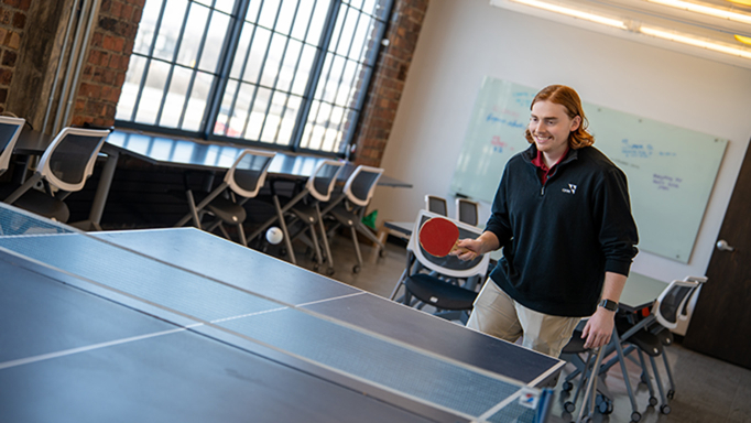 Employee wearing logoed jacket playing ping pong in the office. 
