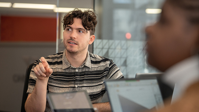 Male employee wearing a striped shirt sitting at his laptop talking to a coworker