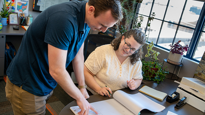 Employee who is standing points to a book on a desk, while woman looks on